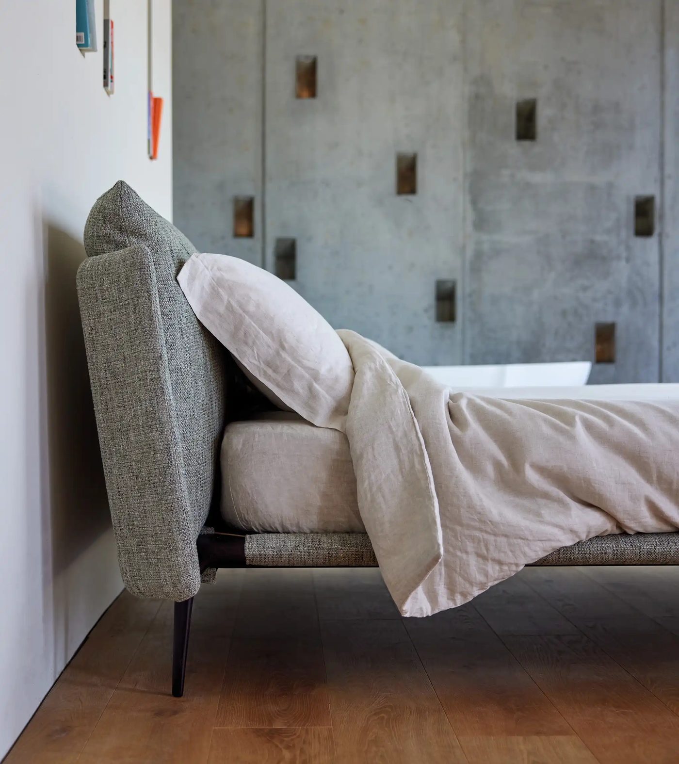 Letto Alto con testiera imbottita in tessuto grigio e biancheria in lino, visto di lato