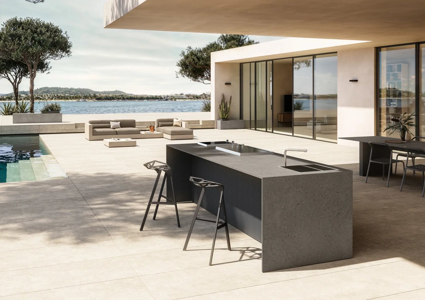 Cucina da esterno Pilan con isola in gres effetto pietra e sgabelli in metallo, su una terrazza affacciata sul mare con piscina e divani outdoor